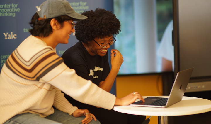  Two people sit at a small table, smiling and looking at a laptop.