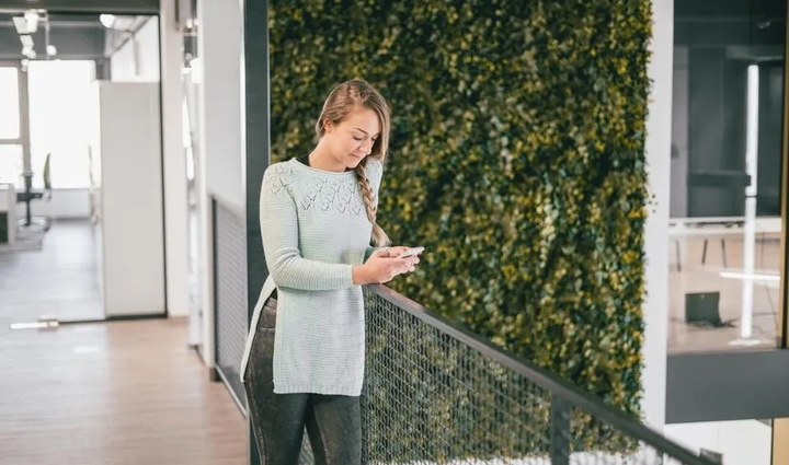 A woman in an office building checks her phone while standing, surrounded by modern architecture and bright lighting. 