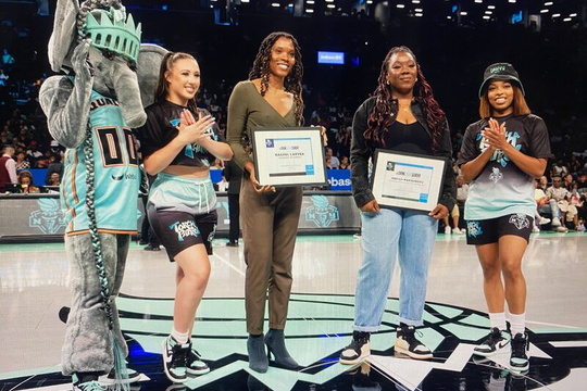 Three women stand on a basketball court holding framed certificates, flanked by a person in a Statue of Liberty costume and another in a team jersey and hat.