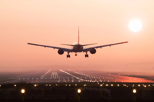 An airplane is landing on a runway at sunset, with the sun low on the horizon and runway lights illuminated.