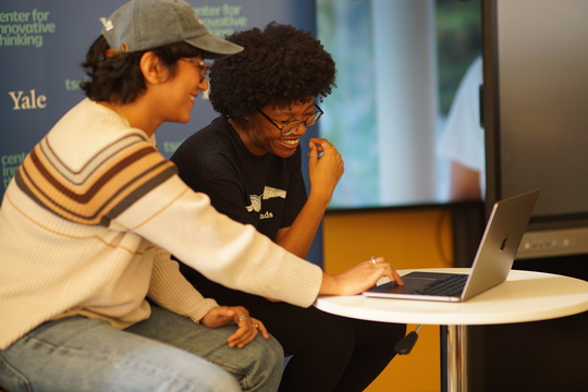  Two people sit at a small table, smiling and looking at a laptop.