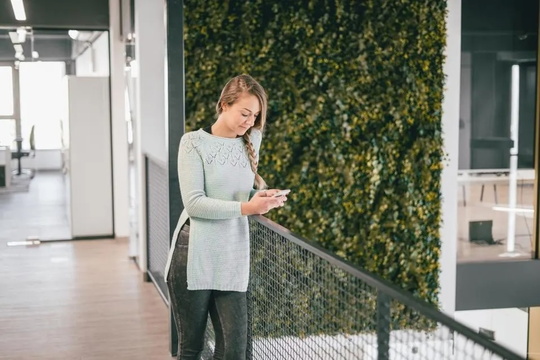 A woman in an office building checks her phone while standing, surrounded by modern architecture and bright lighting. 